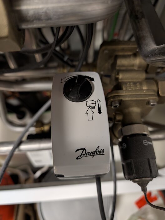 Danfoss reglerventil på fjärrvärmecentral, visande medsols rotationssymbol och omgivande rör och kablar i ett trångt utrymme.
