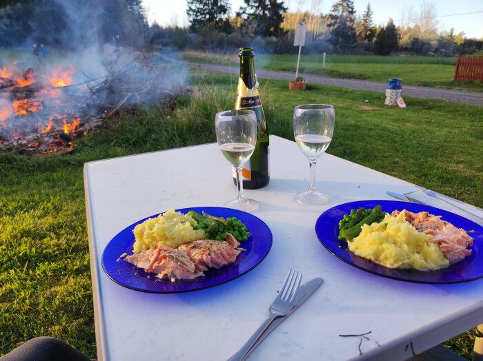 Tallrikar med lax, potatismos och sparris utomhus, två vinglas och champagneflaska på bord, brinnande brasa i bakgrunden.