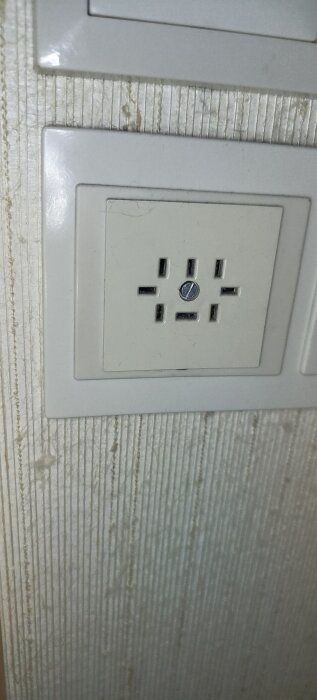 Gammalt vägguttag för intercomsystem, med flera spår runt en centrerad skruv, monterat på en randig tapet.