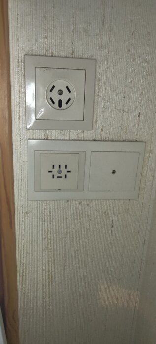 Dubbla vägguttag i beige, troligen del av gammalt intercomsystem, bredvid telefonuttag, mot randig vägg.