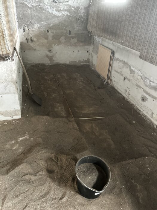 Källarrum med borttagen golvbeläggning, synlig sand och betongväggar, en spade och en hink fylld med sand på golvet.