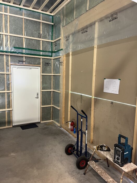 Isolerat rum med plastskikt, 2x2-reglar, och lösull i taket. Dörren, en pirra, brandsläckare och måttband synliga. Stege och radio på golvet.
