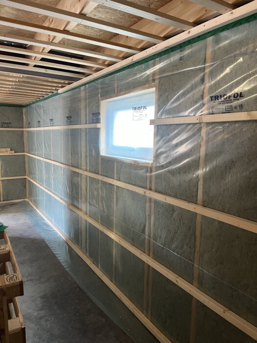 Isolerad vägg med plastfolie och träreglar, pågående byggprojekt med eldragning syns genom ett fönster.