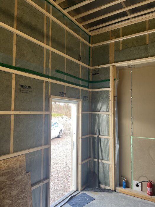 Isolerade väggar med plast och träreglar. Dörröppning visar grusplan med bil. Eldragning pågår. Verktyg och brandredskap vid väggen.