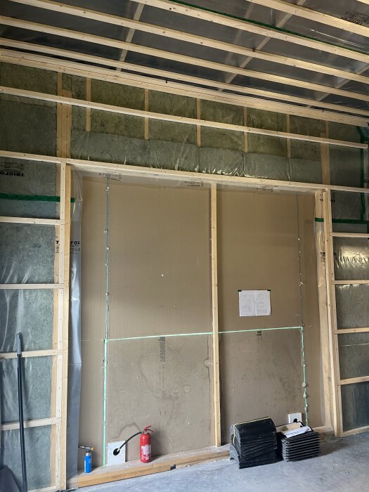 Väggar med isolering och plastfolie, synlig trästomme, utrustning på golvet, inklusive brandsläckare och byggplaner på väggen.