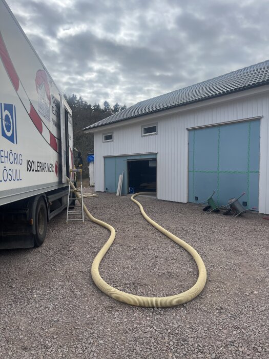 Lastbil med slang för lösullsisolering vid vit byggnad.