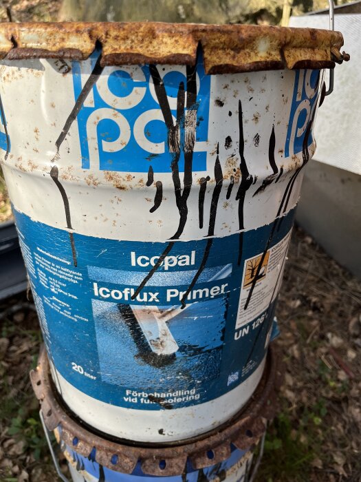 Rostig burk med Icopal Icoflux Primer, placerad på marken i naturlig miljö.