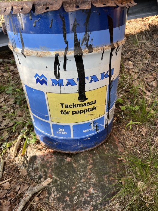 Blå plåtburk med märkning "Täckmassa för papptak", stående på marken, delvis täckt av rost och svart spill.