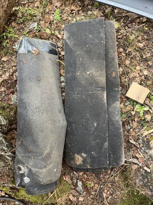 Rullar av gammal takpapp ligger på marken bland löv och gräs, delvis täckta av smuts och kvistar.