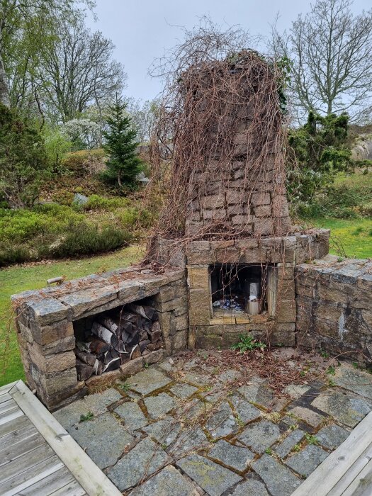 Gammal stengrill med murbruksskador, övervuxen av murgröna, stående på stenlagt underlag, omgiven av buskar och träd i bakgrunden.