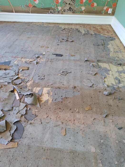Gammalt plankgolv under rivning med kvarvarande papp/filtlager och synliga spår av rivet plastgolv och små spikar. Blommig tapet i bakgrunden.