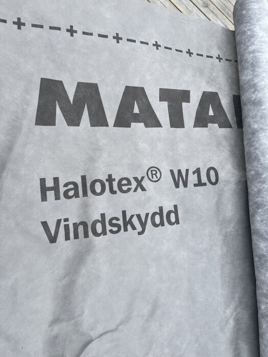 Rulle av Halotex vindskyddsmaterial ligger på träyta, med texten "Halotex W10 Vindskydd" synlig.