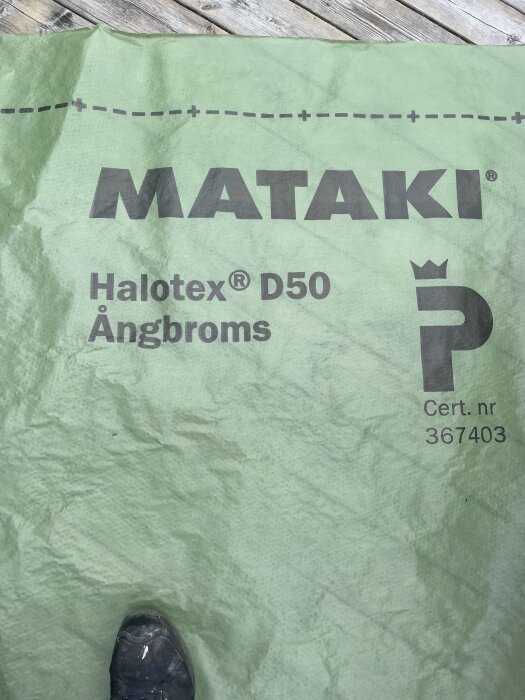 Grön byggplast med texten "MATAKI Halotex D50 Ångbroms" på en träyta, en del av en sko syns.