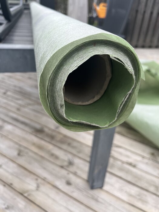 Grön byggpappersrulle delvis upprullad på en metallställning, på träunderlag utomhus.