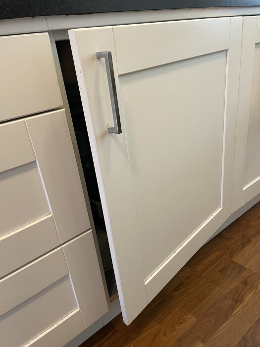 Vit kökslucka med silverhandtag som står öppen några centimeter, del av diskbänk. Luckan ser ut att inte kunna hålla sig stängd.