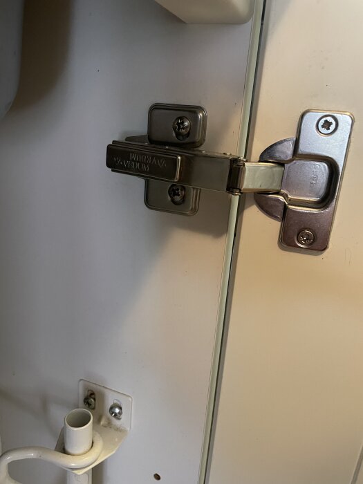 Närbild på gångjärn på en kökslucka under diskhon, märkt Vedum. Luckan har problem att stänga helt.