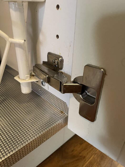 Gångjärn på kökslucka under diskho, delvis öppen, med synlig montering och intilliggande rör.