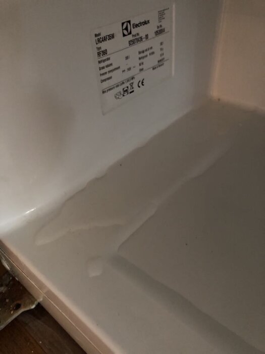 Kylskåpsinteriör med vattenläckage under en vit hylla och modellnummer synligt på en etikett på väggen.