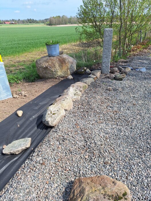 Stenar placerade på markduk vid en grusuppfart, med ett fält och några träd i bakgrunden. En hink står på en stor sten.