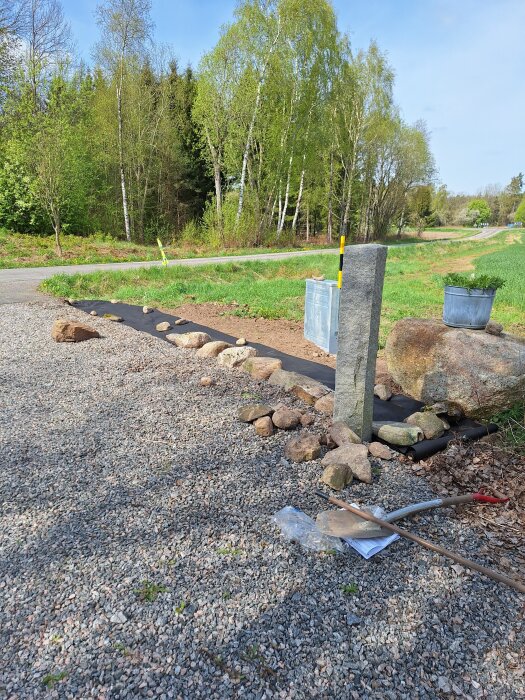Grusplan med rensade stenar och redskap, omgiven av träd i soligt landskap.