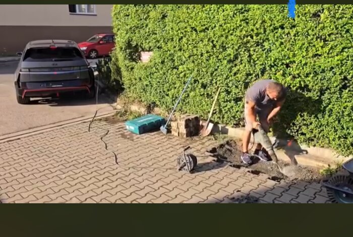Man häller betong på stenlagd uppfart bredvid häck; elbil och verktyg i bakgrunden.