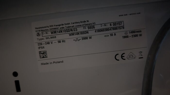 Etikett på tvättmaskin med produktnummer, serienummer, tillverkningsland och tekniska specifikationer som spänning och effekt.