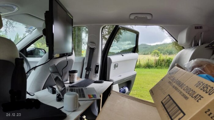 Arbetsplats i bil: laptop, skärm, kaffemugg och kartonger, parkerad i ett grönt fält med öppna dörrar, natur i bakgrunden.
