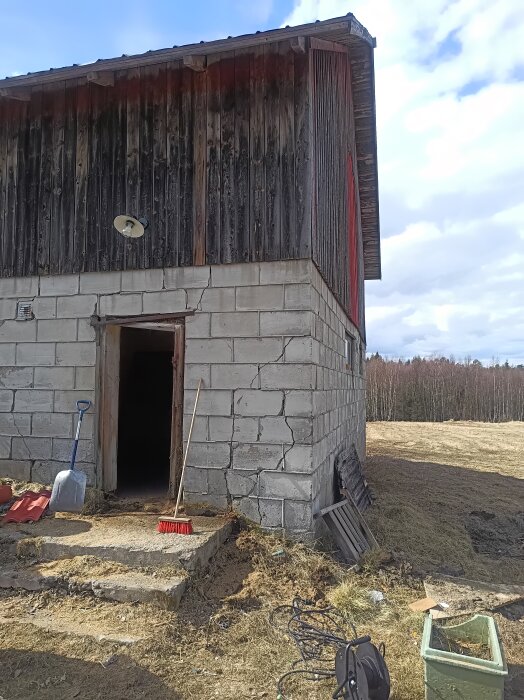 Sprickor i lättbetongvägg på gammal ladugård, träpanel ovanpå, verktyg vid entrén, rural miljö i bakgrunden.