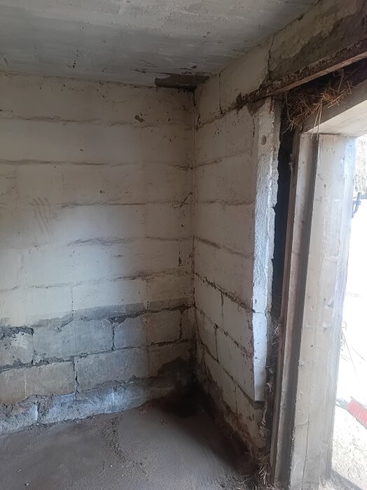 Sprucken yttervägg i lättbetong med lös karm och en synlig spricka vid hörn, med delar av timmer och bråte vid en dörröppning.