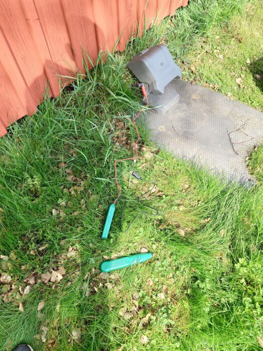 Elektroniska verktyg och en laddstation för robotgräsklippare på grönt gräs vid en röd vägg. Kabel draget på marken.