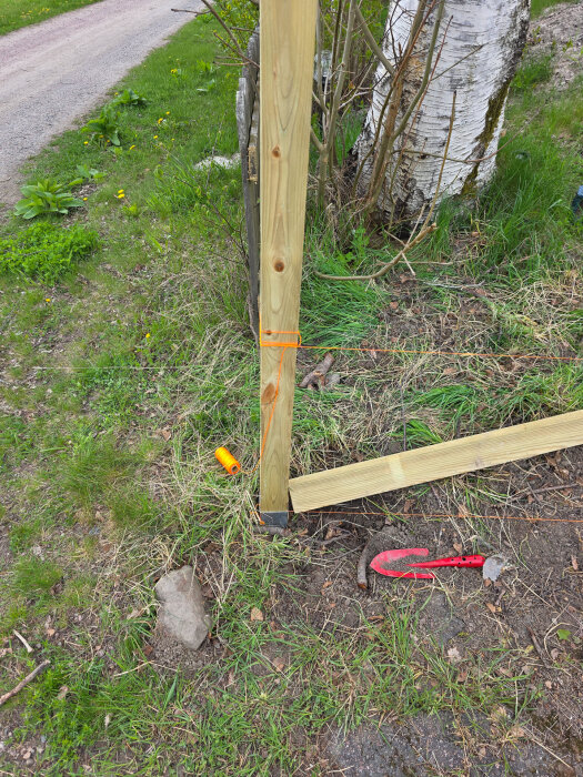 Trästolpe monterad i ett stolpspjut bredvid en björk, orange murarsnöre, ett nedböjt verktyg och ett rör på marken, gräs och gångväg i bakgrunden.