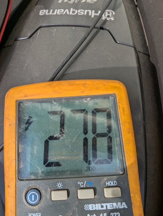 Multimeter visar 27.8V bredvid Husqvarna-robotgräsklippare, använd för att mäta laddningsspänning och diagnostisera problem med klipparens drift.