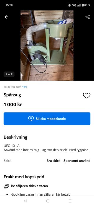Grön spånsug med slang, stående inomhus på ett trägolv nära en vägg av träplankor.