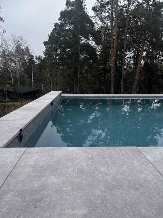 Pool med blått vatten omgiven av grå bänkskivor och skog i bakgrunden.
