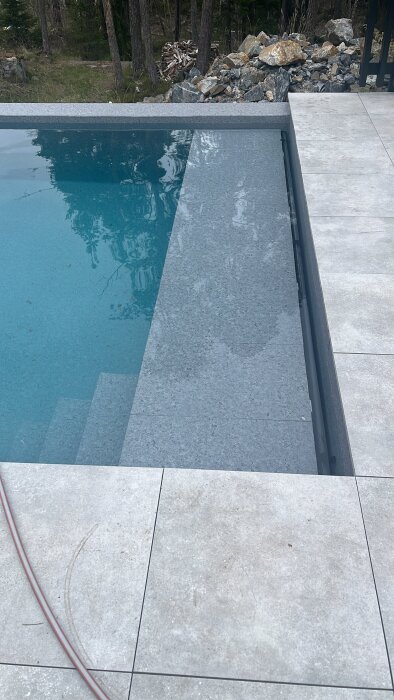 Pool med nylagt kakel och klart blått vatten, omgiven av stenhällar, med träd och stenar i bakgrunden.
