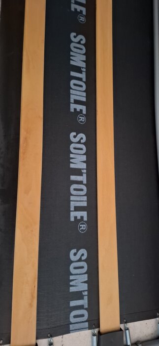 Närbild av bäddsoffans ribbor och tyg med texten "SOM'TOILE" synlig på duken.