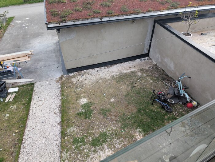 Innergård med gräsyta och grusgång, cyklar och en röd boll ligger mot en vägg. Byggnad med takvegetation i bakgrunden.