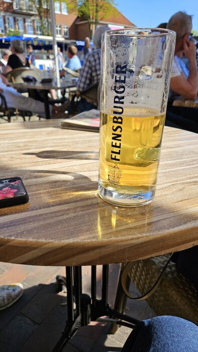 Ett glas Flensburger-öl på ett bord utomhus vid Heiligenhafens 1:a majfirande. Människor syns sitta vid bord i bakgrunden.