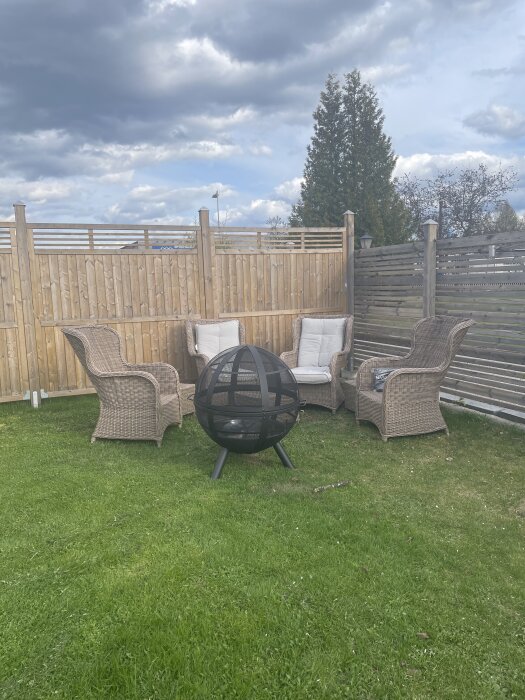 Trädgårdsplats med flätade stolar, en eldkorg och ett trästaket i bakgrunden under molnig himmel.
