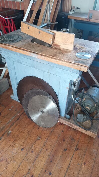 Gammaldags snickeribord med sågblad och verktyg på slitet trägolv i en snickeriverkstad, omgiven av träbitar och utrustning.