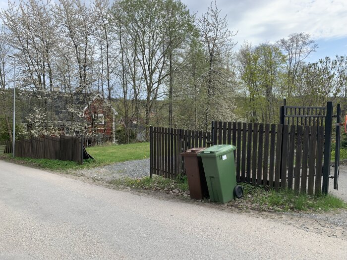Trästaket vid en väg med soptunnor framför, omgivna av gröna träd och en röd byggnad i bakgrunden.