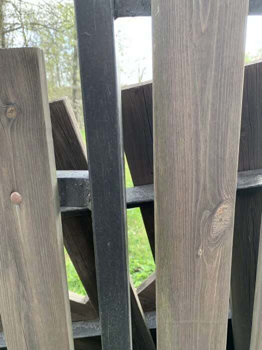 Trästaket med störar och metallstolpe, omgiven av grönska.