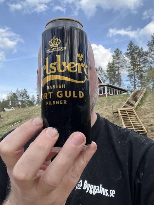 En person håller en burk Carlsberg Sort Guld Pilsner framför ansiktet utomhus, med ett hus och trappa i bakgrunden.