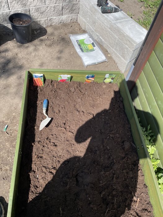 Planteringslåda med jord och en handspade, fröpåsar på kanten. Verktyg och kruka i bakgrunden, bredvid en stenmur och grön plankvägg.