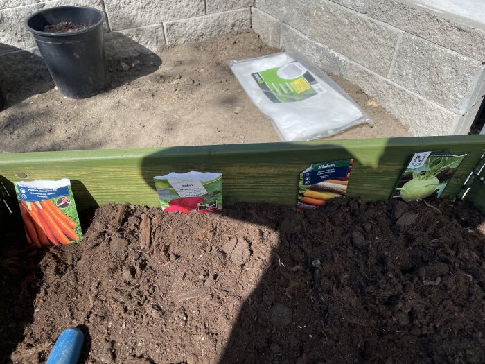Fröpåsar av morötter, rädisor och kålrot i en odlingslåda, med jord färdig för plantering. En plastkruka och säck syns i bakgrunden.