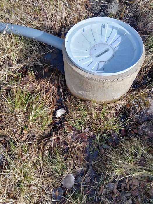 Cementbrunn med plastlock, omgiven av fuktig mark och gräs, visar vattenläckage vid skarv. En grå ventilationsrör sträcker sig från brunnens topp.