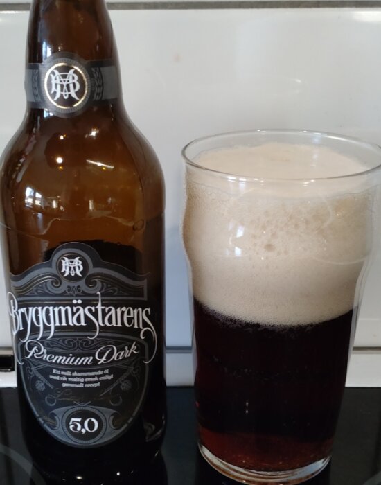 Flaska och glas med mörk lageröl, etikett visar "Bryggmästarens Premium Dark", bredvid ett glas med skumtäckt, mörkt öl.