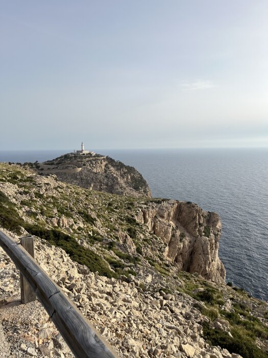 Fyrtorn på en klippa vid havet, omgiven av gröna buskar och stenar, med en räcke i förgrunden och en klar himmel ovanför.