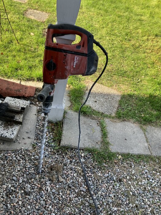 Röd borrmaskin med spiralborr, stående mot en träpelare på en grusad yta i en trädgårdsmiljö.
