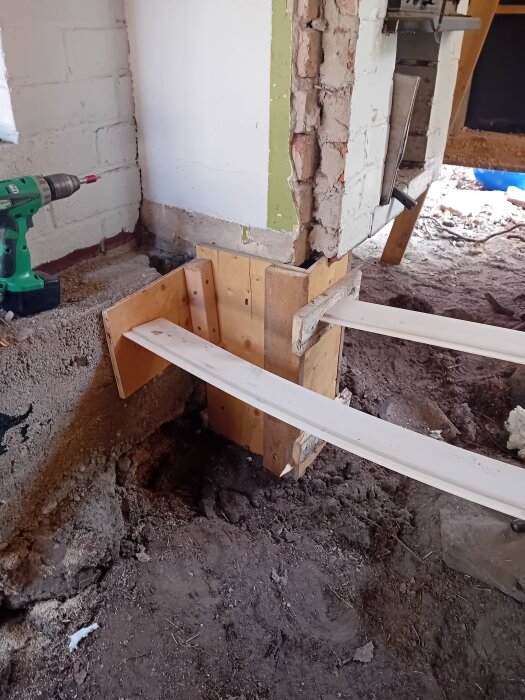 Formar för gjutning av sulor vid grundarbete, visande tegelmur och träformar med borrmaskin liggande på marken.
